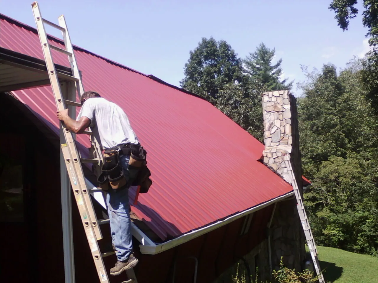 Roofer on a ladder beside a red metal roof and stone chimney — Soffit & Fascia Repair by Roofing Brothers in Talmage